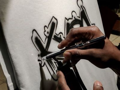 A hand holds an airbrush gun painting block lettering onto a white shirt.