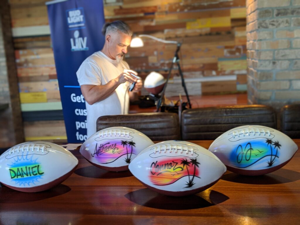 custom airbrushed footballs sit in the foreground while an airbrush artist paints another football in the background