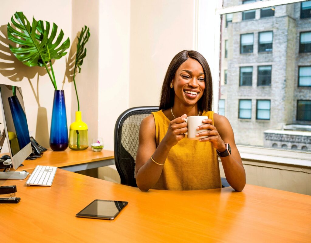 a lady sits at a large desk holding a mug of coffee in front of a large window looking at another large building next door - The How and Why of Starting a Business in Your Local Community