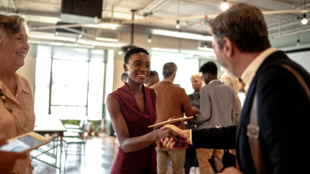 a man and woman shake hands in a large office with other people standing and socializing in the background - DIY MARKETING: How to promote your one-person business