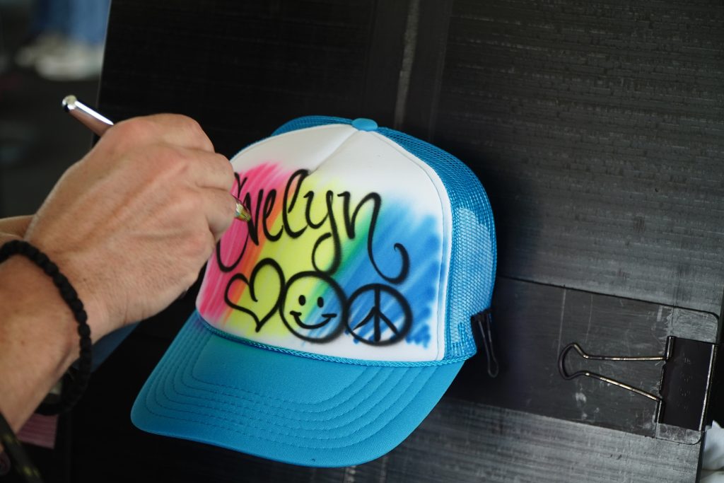 a person holds an airbrush gun painting a blue trucker hat with the name Evelyn on it.
