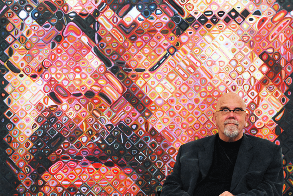 Artist seated in front of large Chuck Close-style photorealistic grid portrait artwork, demonstrating the expensive airbrush artwork technique that made Close famous for million-dollar paintings