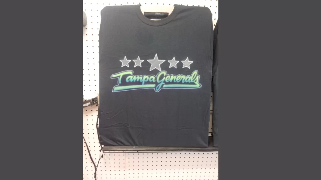 A black t-shirt with the words "Tampa Generals" and star designs airbrushed in vibrant colors. The image demonstrates the visual appeal of airbrushed designs on dark fabrics, which are not ideal for events due to the extensive priming and drying time required.